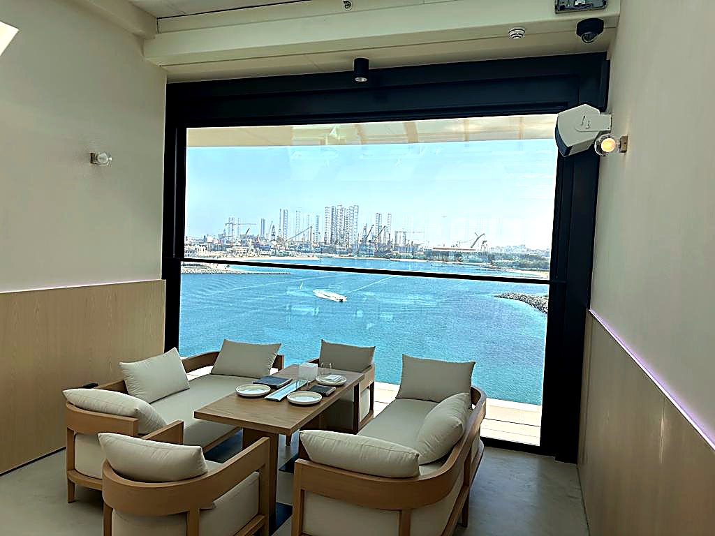 Lounge chairs and low tables beside full height windows overlooking the water